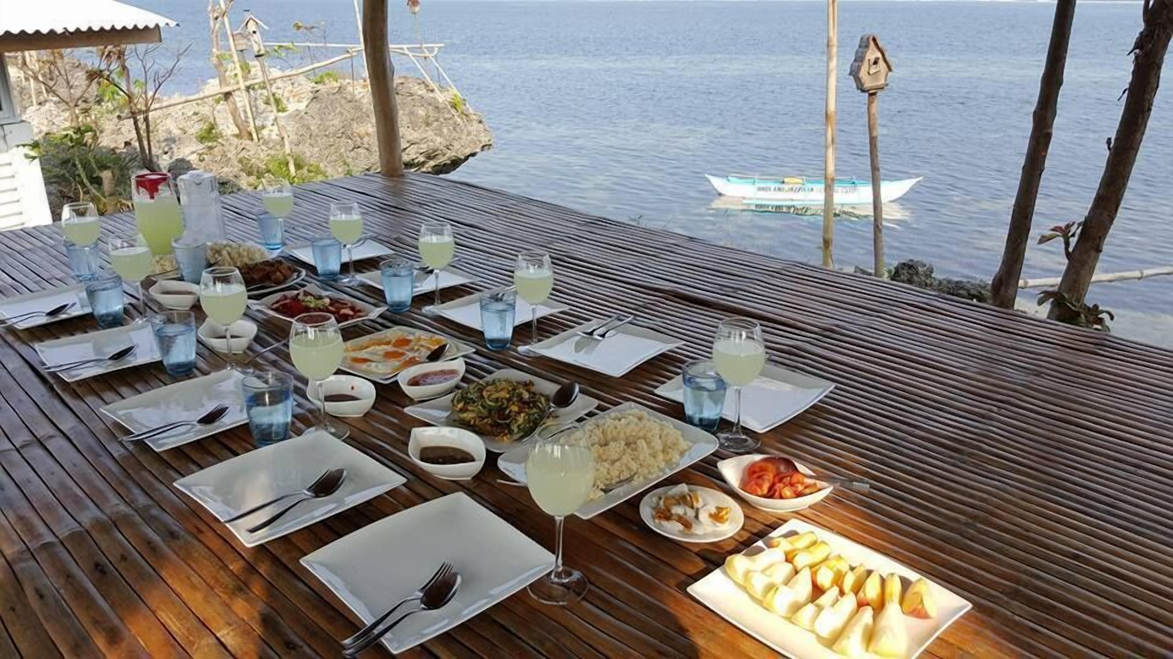 A wooden dining table set with plates, cutlery, and drinks, overlooking a calm sea with a boat in the distance.