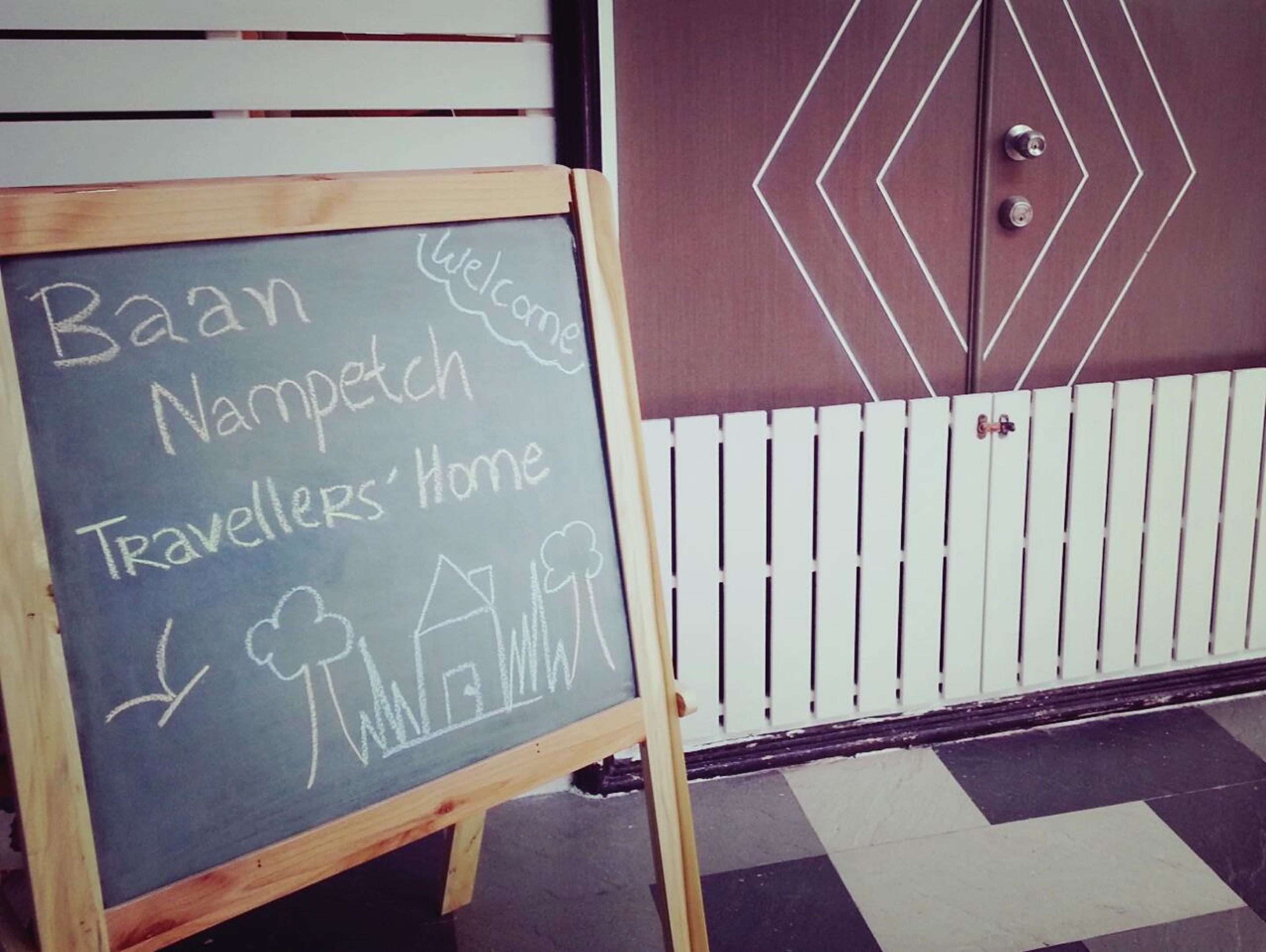 A chalkboard sign with 'Baan Nampetch Travellers' Home' written on it, placed in front of a door with a geometric design and a white picket fence.