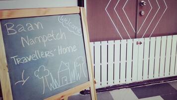 A chalkboard sign with 'Baan Nampetch Travellers' Home' written on it, placed in front of a door with a geometric design and a white picket fence.