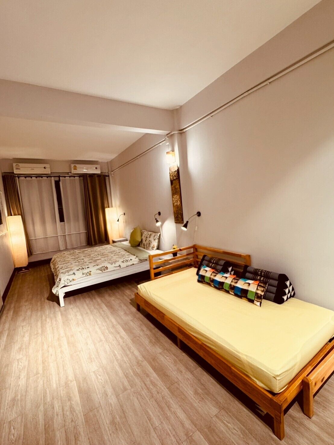 A bedroom with a wooden bed, a smaller bed, a bedside table with a lamp, and a wall-mounted mirror.