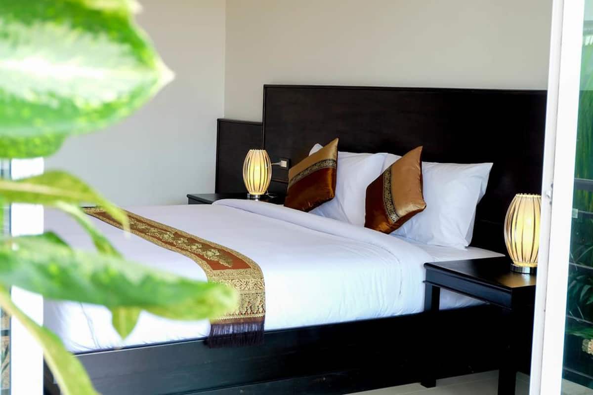 Een net opgemaakt bed met witte lakens en twee decoratieve kussens, een donker hoofdbord en nachtlampjes op een nachtkastje.