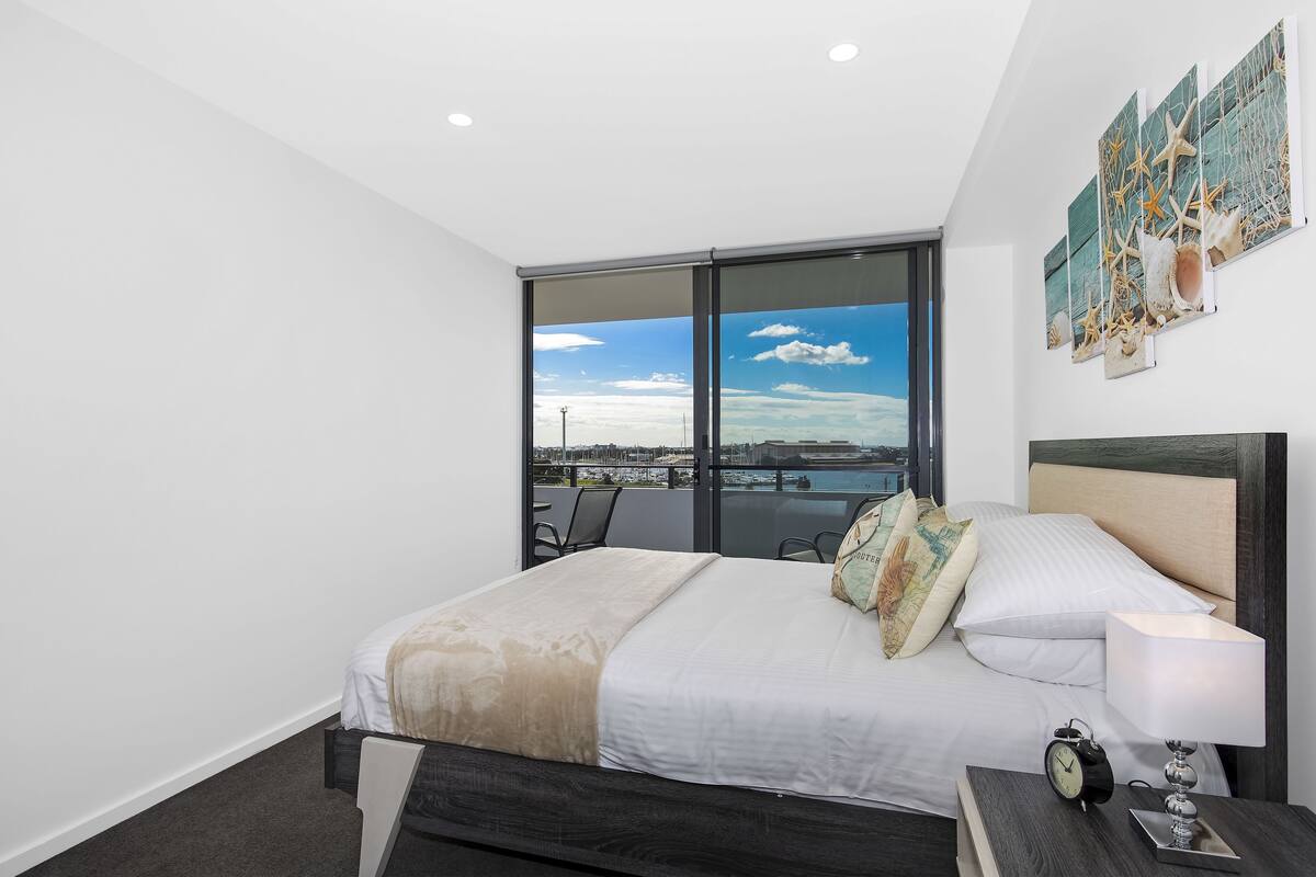 A modern hotel room with a large bed, a nightstand, a lamp, and a view of the city through floor-to-ceiling windows.