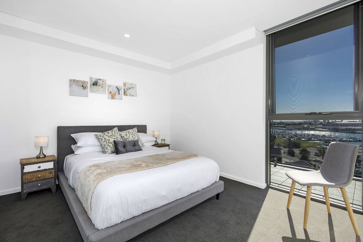 A modern bedroom with a large bed, a nightstand with a lamp, a chair, and a view of a cityscape through a large window.
