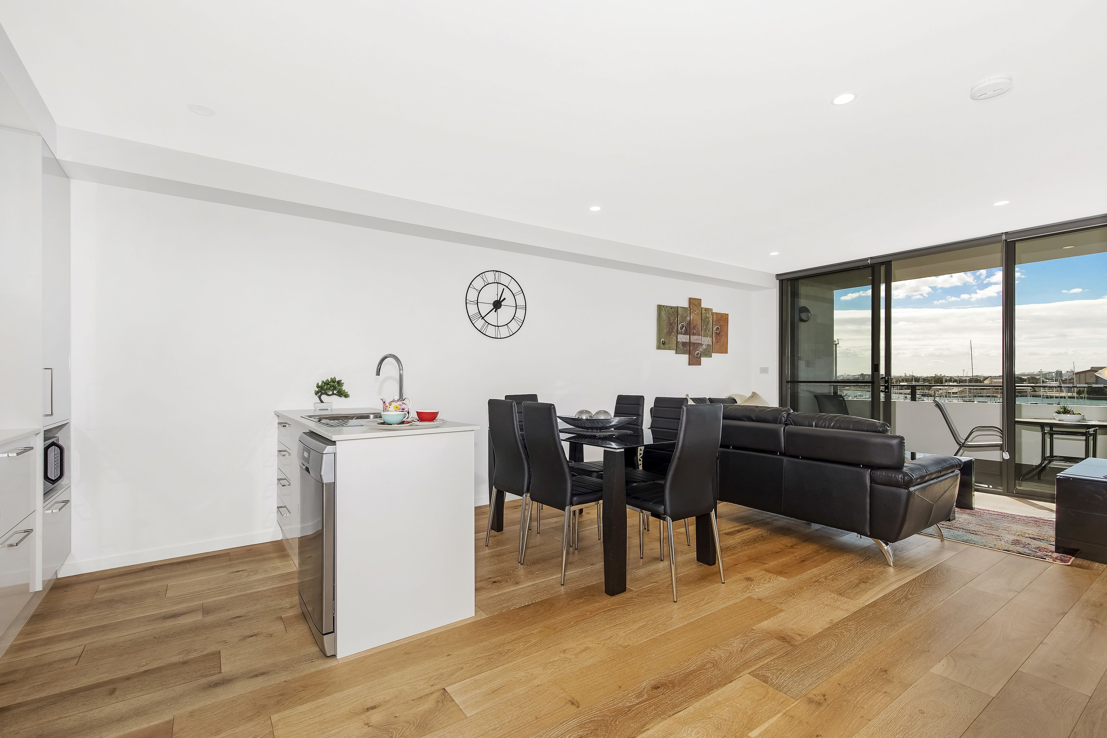 A modern kitchen with a dishwasher, a dining area with a black sofa and chairs, and a large window offering a city view.