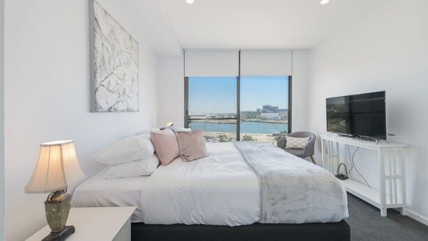 A modern bedroom with a large bed, a TV, and a view of a cityscape through large windows. - Astra Apartments Newcastle West (Newcastle West)