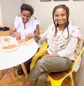Deux personnes sont assises autour d’une table ronde, devant des assiettes de nourriture, dans une pièce dont le sol est en bois et où l’on aperçoit un mur sobre à l’arrière-plan.