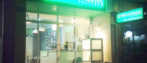 A MicroHostel with a green neon sign, glass doors, and a visible reception area with a counter and chairs outside.