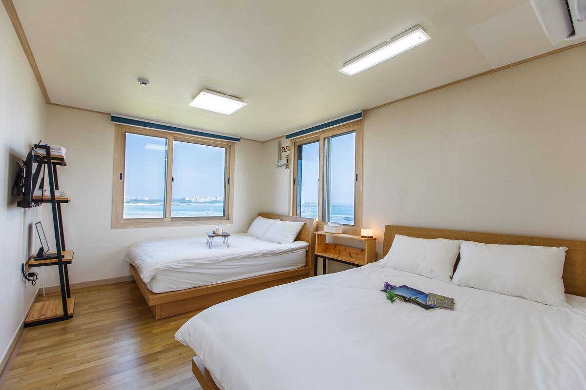 A hotel room with two beds, wooden flooring, a desk, and large windows offering a view of the sea.