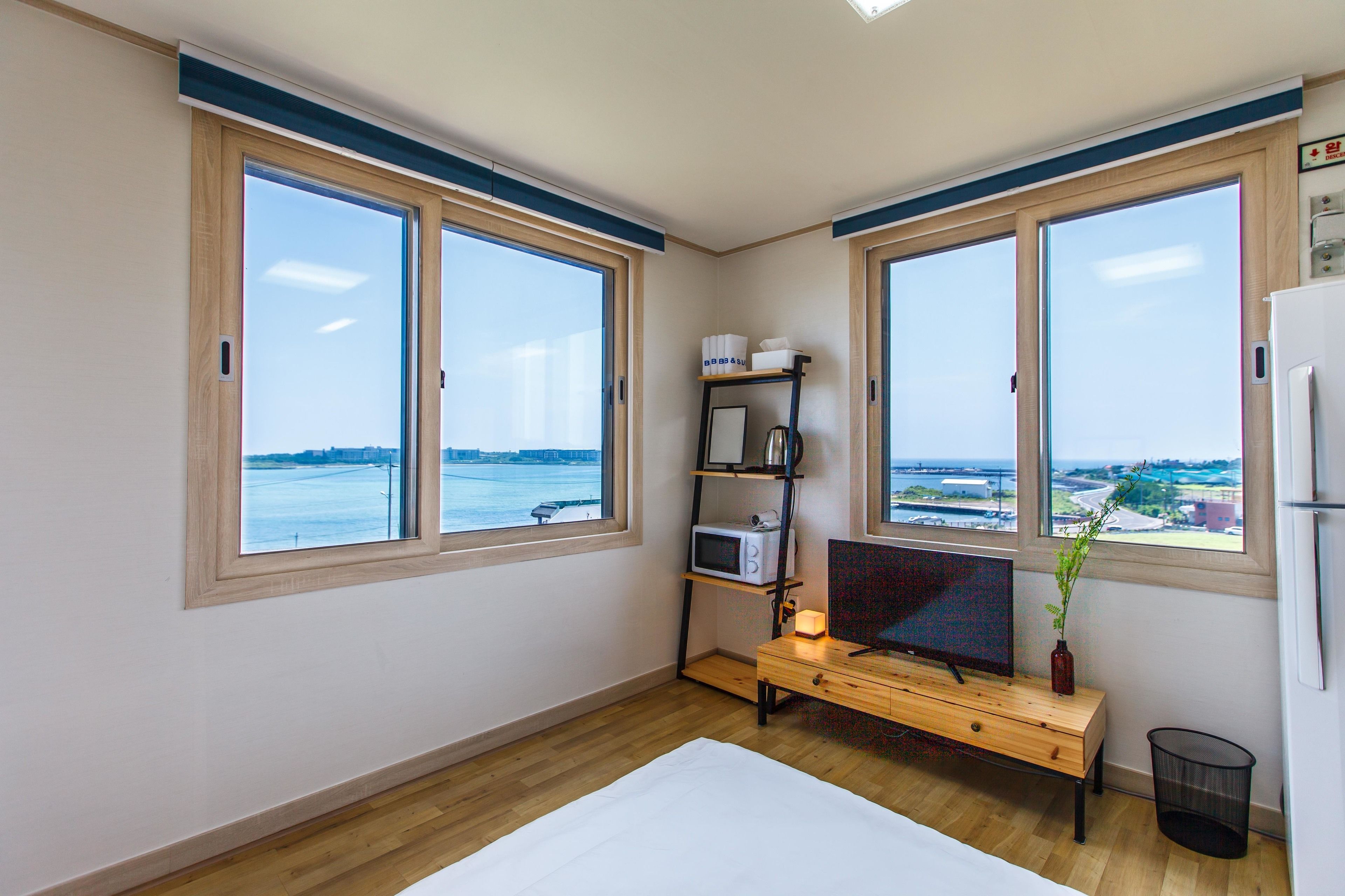 A room with two large windows, a wooden desk, a television, a small shelf with items, and a view of a body of water.