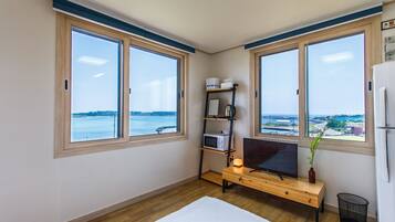A room with two large windows, a wooden desk, a television, a small shelf with items, and a view of a body of water.