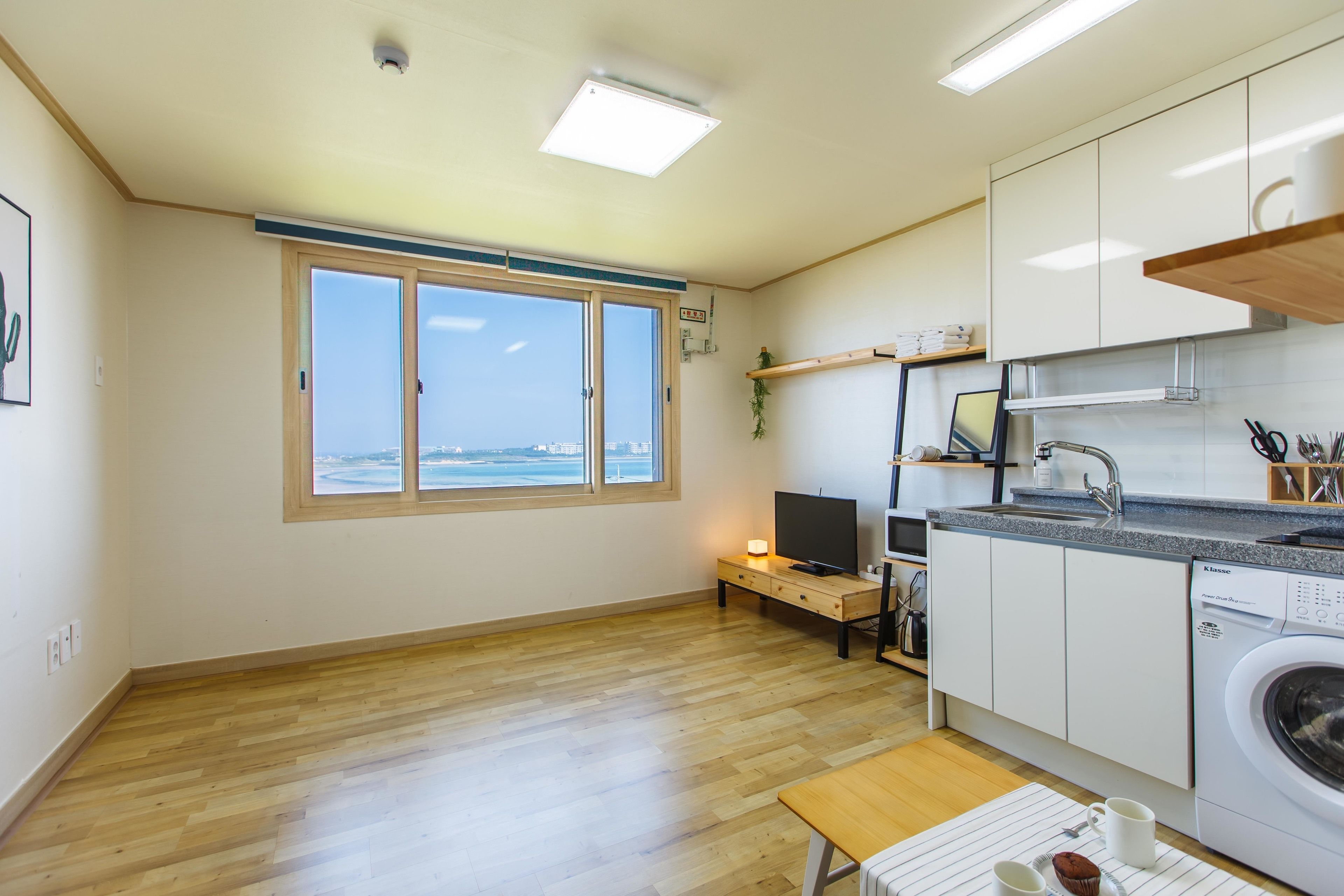 A modern kitchen with wooden flooring, a built-in dishwasher, and a large window overlooking the sea.