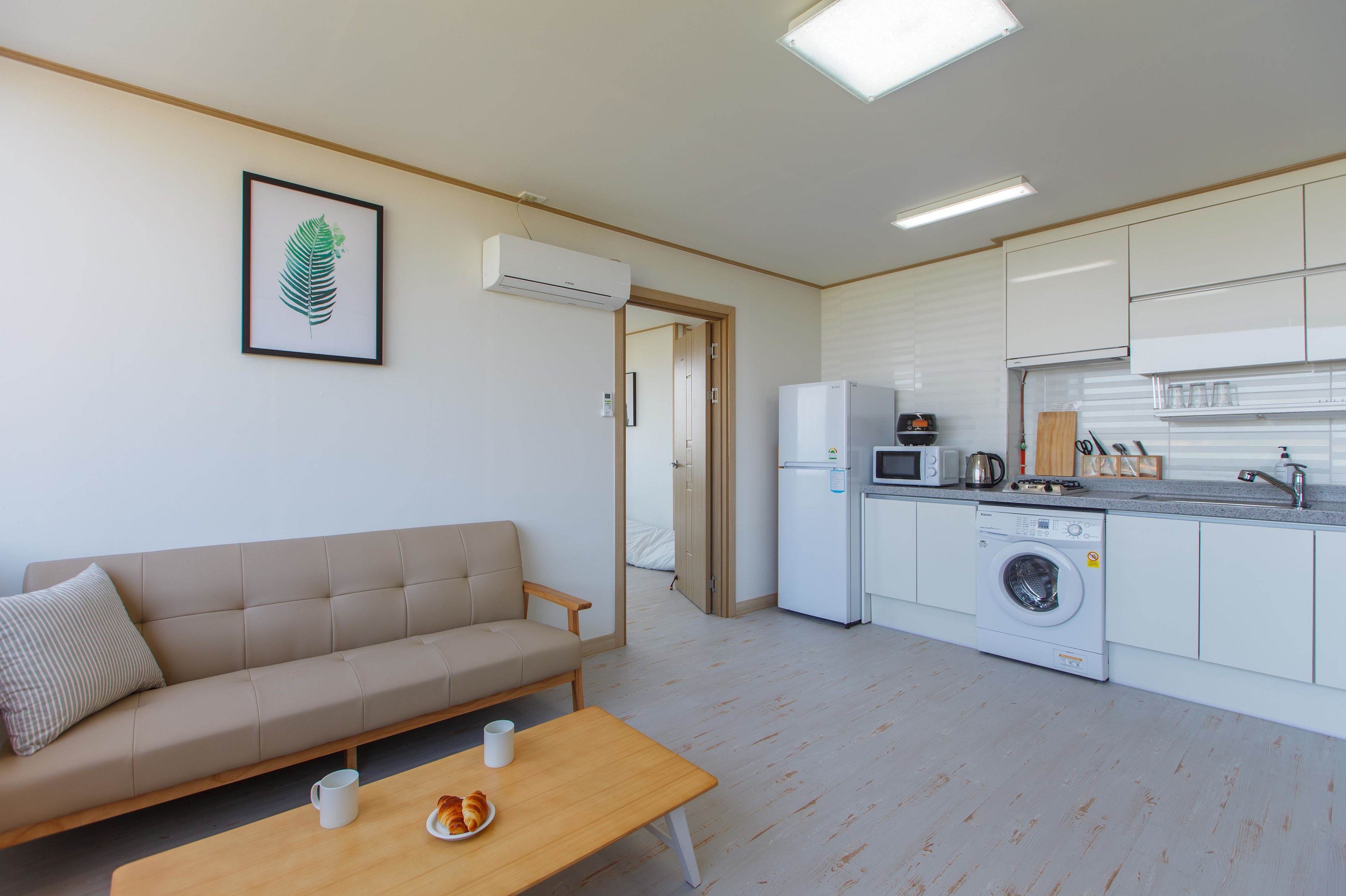 A compact living space with a sofa, a small table, a washing machine, a microwave, and a kitchenette.