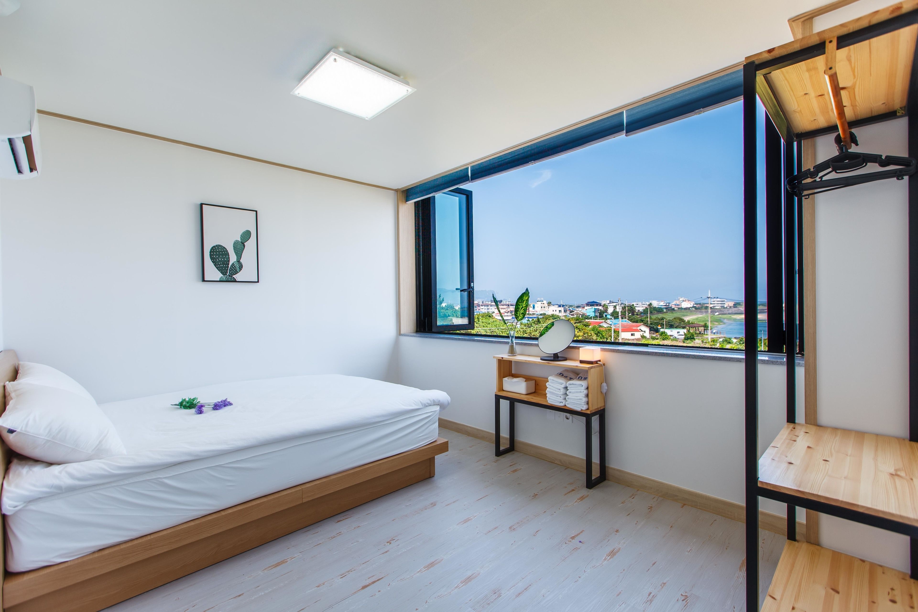 Quarto minimalista com cama, mesa de cabeceira com espelho e vista da cidade através de uma grande janela.