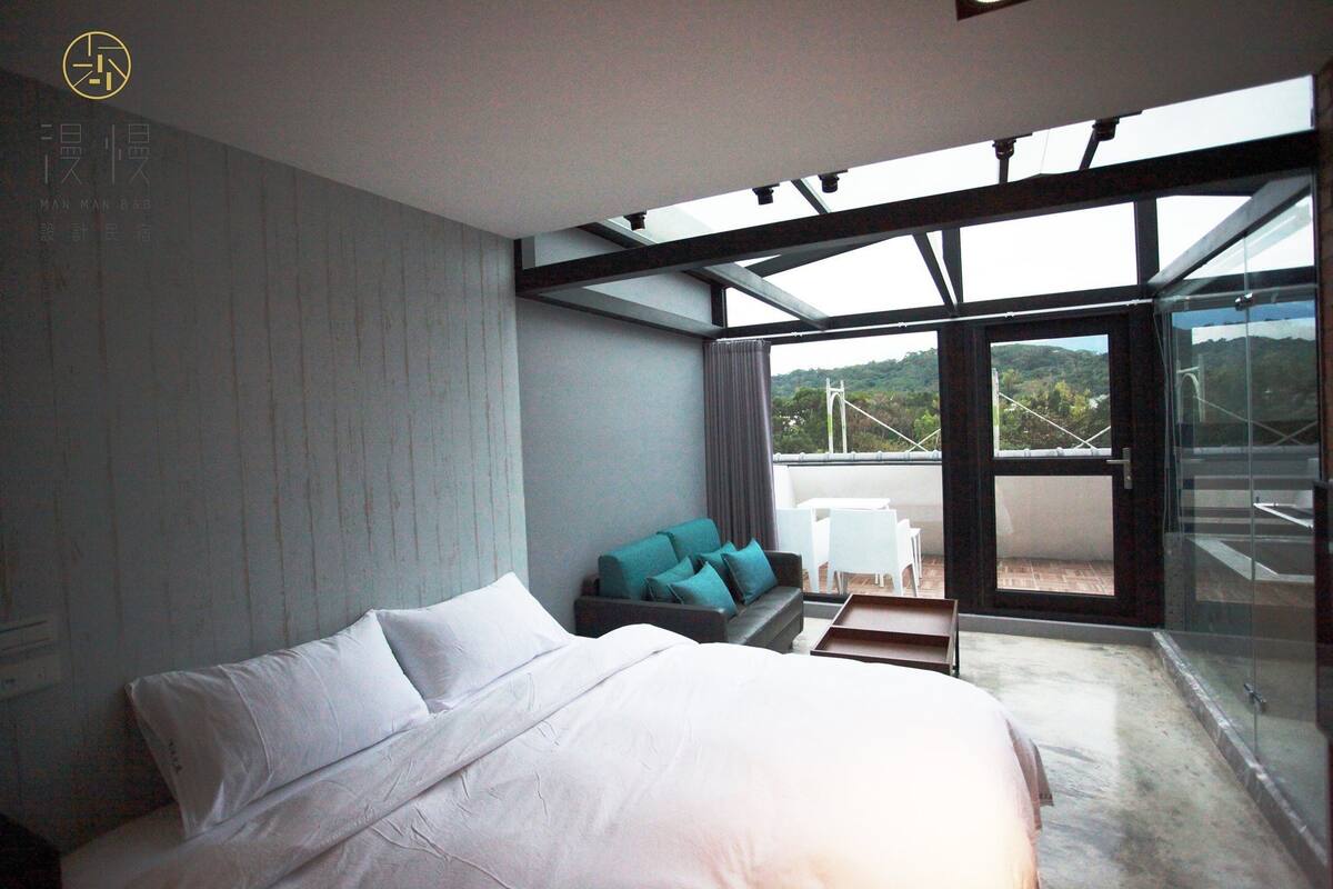 A modern bedroom with a large bed, a sofa, and a glass door leading to a balcony.