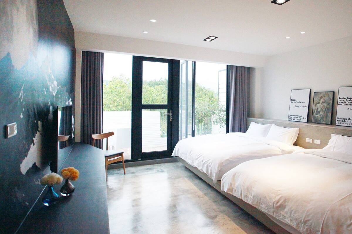 A modern hotel room with a large bed, a desk, and a chair. There are two windows with curtains, and a view of greenery outside.