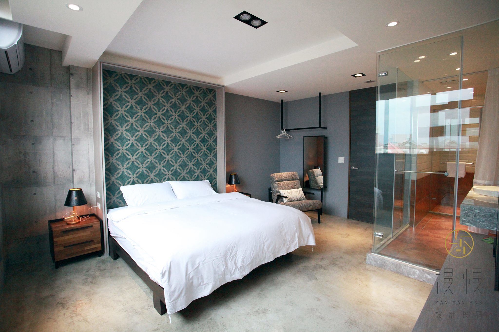 A modern bedroom with a large bed, a nightstand, a chair, a glass shower enclosure, and a patterned wall.
