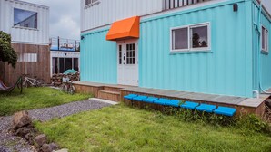 Un bâtiment moderne à la façade turquoise, avec une terrasse en bois dotée de bancs bleus et un vélo garé à l’extérieur.