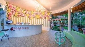 A reception area with a curved counter, a chandelier, and a sign that reads "Reception". There is a seating area with a glass table and a cushioned bench. - Baan Aom Kod Kun Kao (Suan Phueng)
