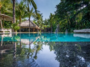 Une piscine aux eaux cristallines dans laquelle se reflètent les palmiers, ainsi qu’un bâtiment à toit de chaume.