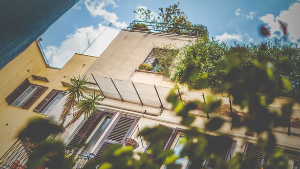 A building with multiple windows, some with shutters, and a balcony with a plant. - Domus Alberti (Rome)