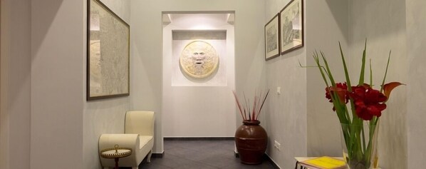 A hallway with a framed artwork on the left, a vase with red flowers on the right, and a chair in the center. - Domus Alberti (Rome)