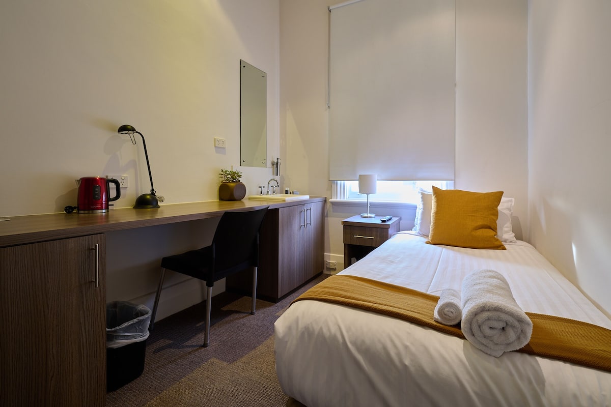 Ett hotellrum med en säng, ett skrivbord med en stol, ett litet bord med en lampa, en röd vattenkokare och ett fönster med en gul kudde.