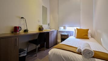 Hotelkamer met een bed, een bureau met een stoel, een tafeltje met een lamp, een rode waterkoker en een raam met een geel kussen.