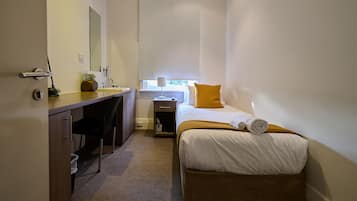 Une chambre d’hôtel avec un lit, un bureau, une chaise, une petite table, une plante, une fenêtre avec un store et un miroir au mur.