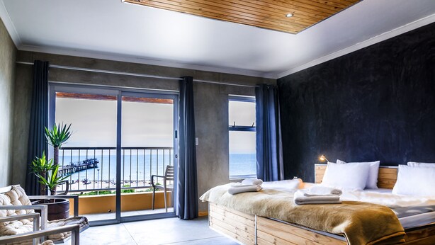A modern hotel room with a large bed, a balcony with a view of the sea, and a wooden ceiling.