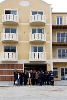 Un edificio de varios pisos con balcones, un grupo de personas de pie en la entrada y un acceso al garaje.