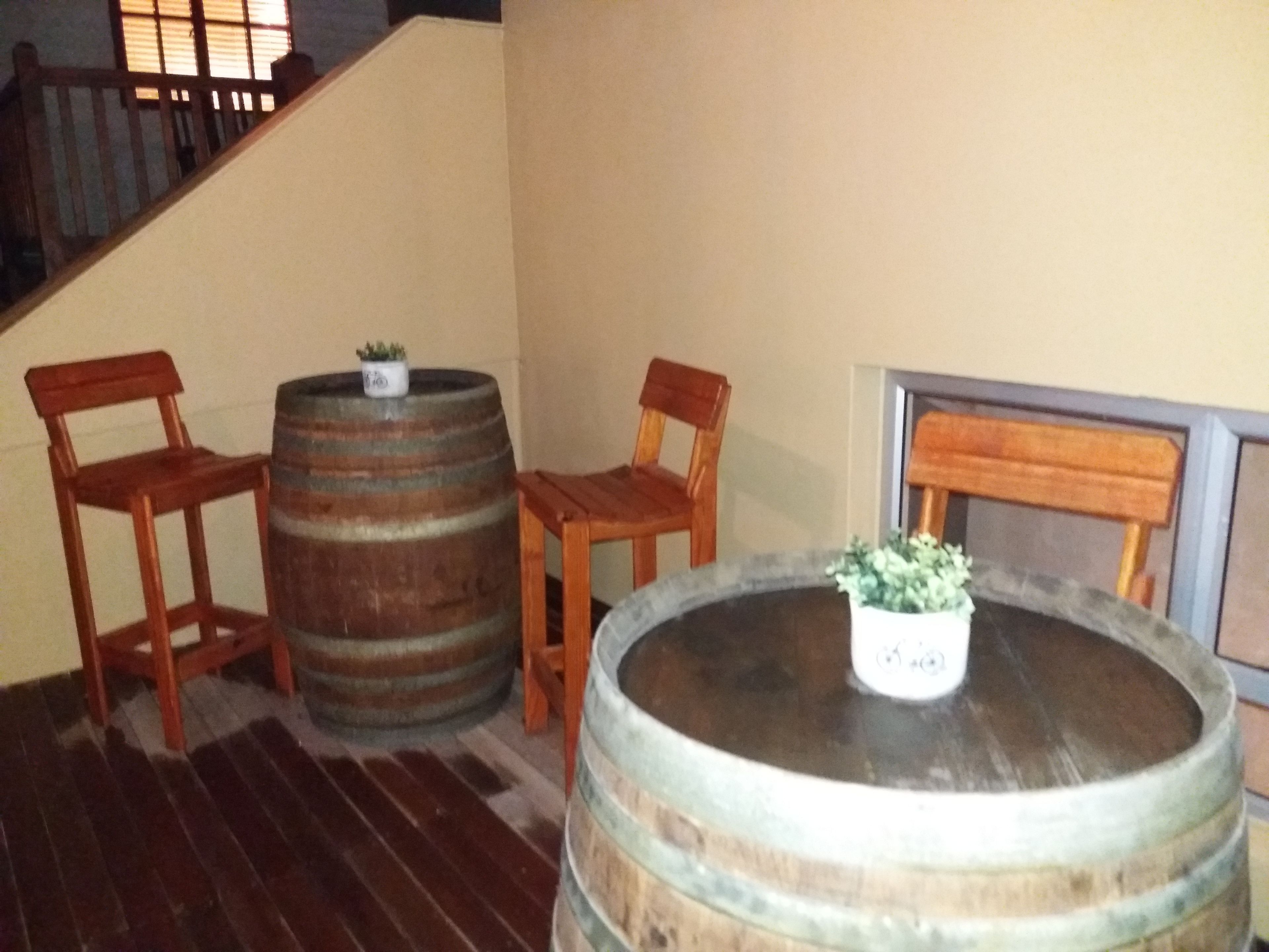 A room with a wooden floor, two wooden chairs, a round wooden table with a potted plant, and two wooden barrels.