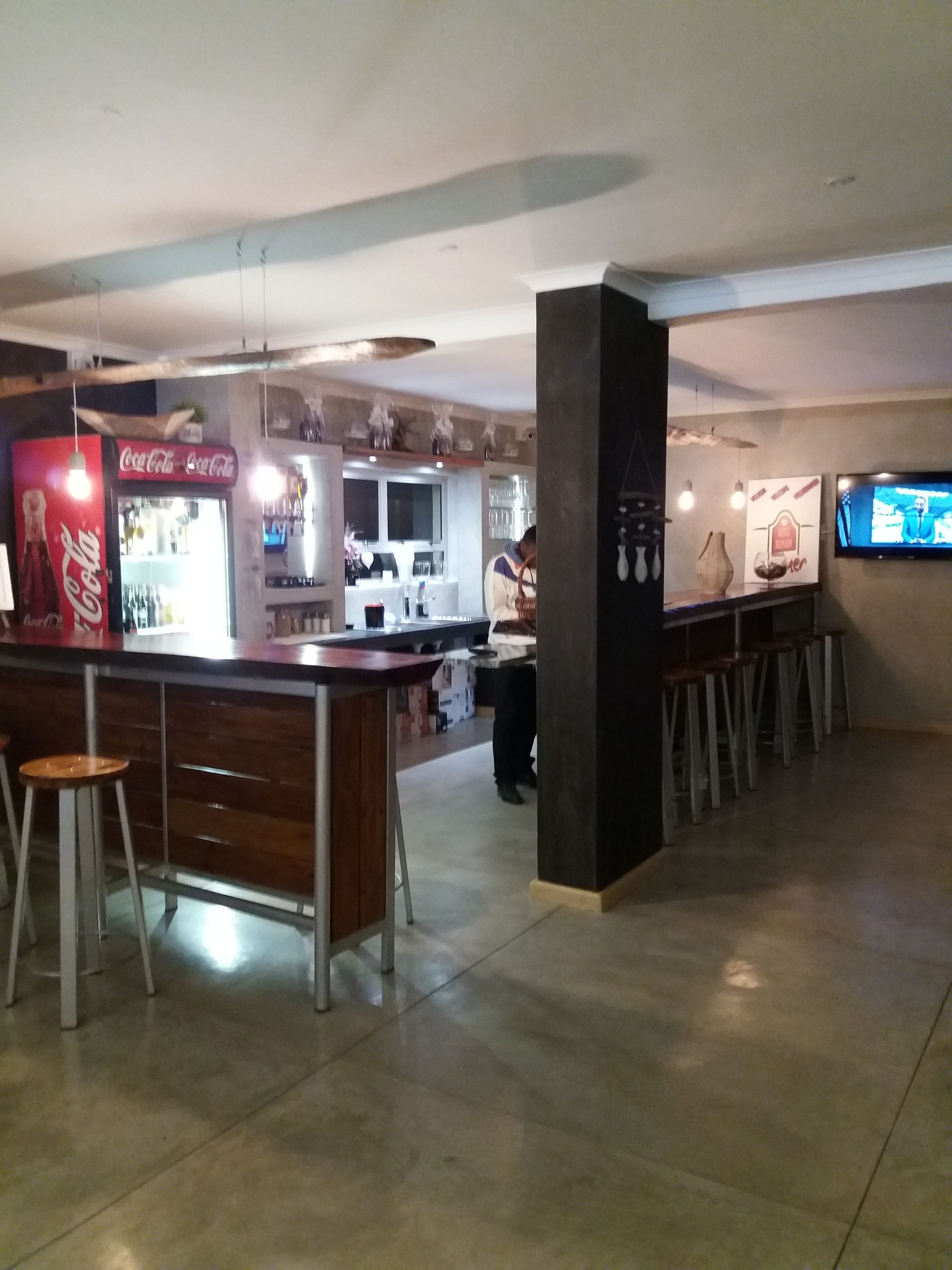 A bar area with stools, a counter, and a Coca-Cola refrigerator. There are hanging lights and a television mounted on the wall.