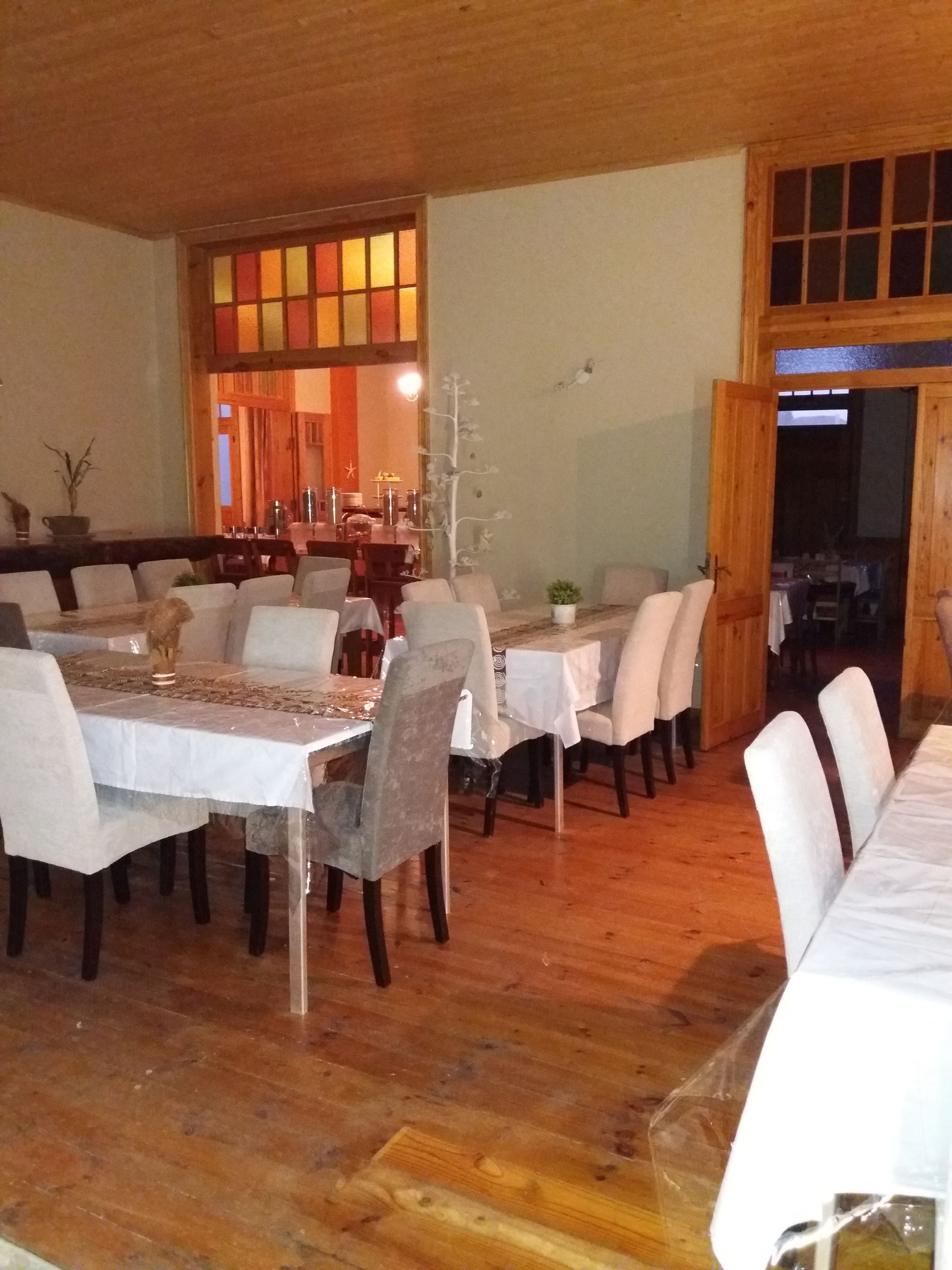 A dining area with multiple tables and chairs, wooden flooring, and a view of a kitchen area through an open doorway.