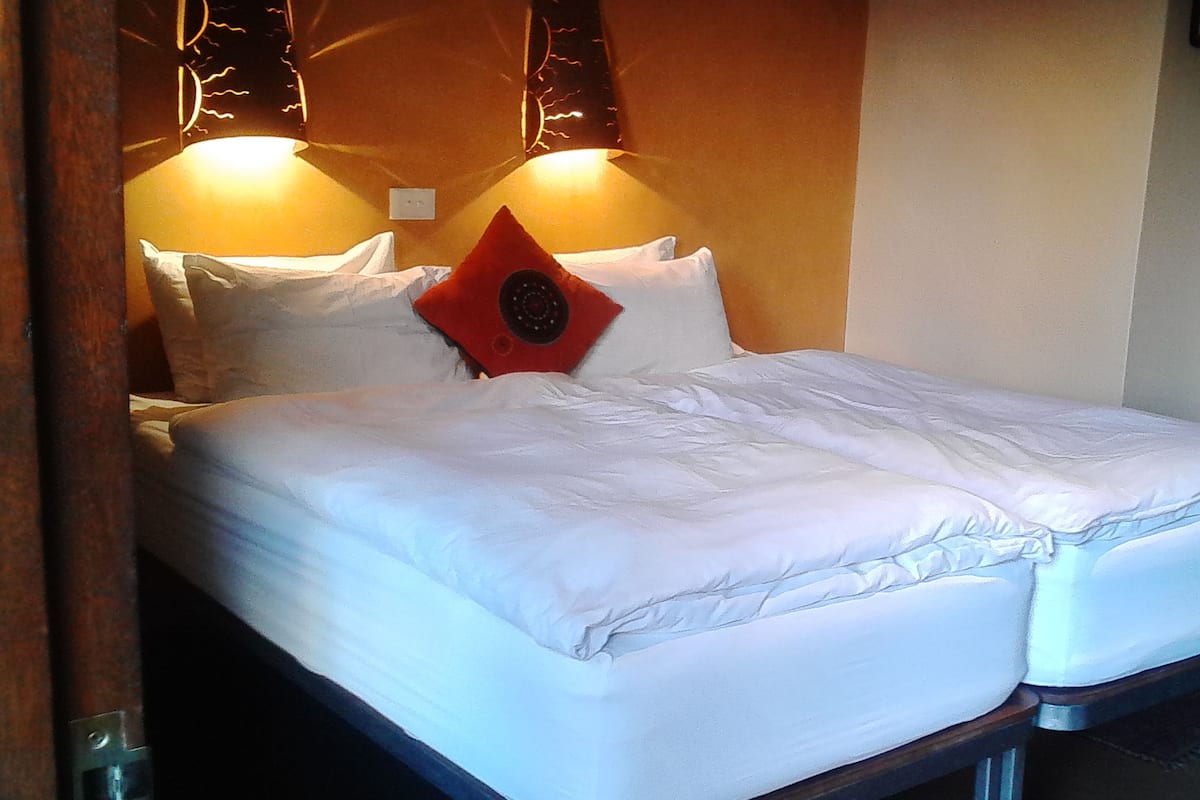 Una cama doble con sábanas blancas, un cojín decorativo rojo y dos lámparas fijadas a la pared.