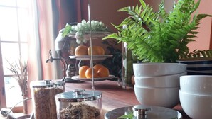 A buffet setup with various food dispensers, including cereal and granola, and a fruit display with oranges and grapes.