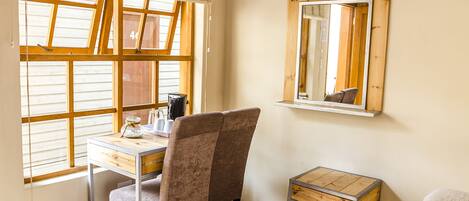 A room with a wooden desk, a chair, a small table, a mirror, and a window with blinds.