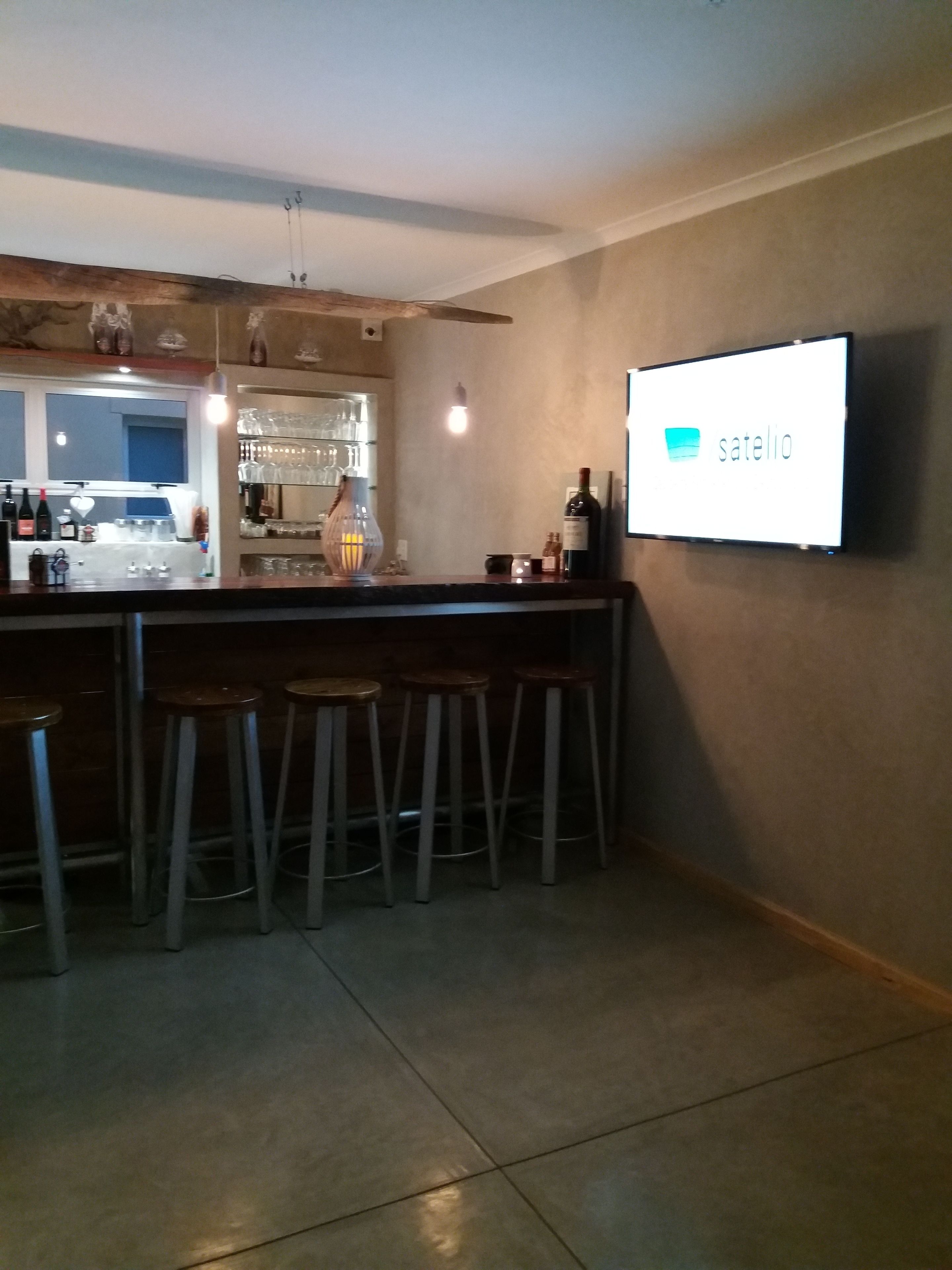 A bar area with stools, a TV screen displaying 'satelio', and a counter with bottles and glasses.