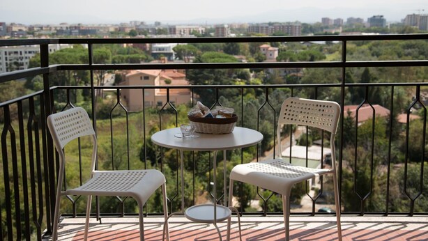A balcony with two white chairs and a small round table, overlooking a cityscape with buildings and greenery. - My Sweet Rome (Rome)