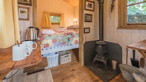 A cozy room with a bed, a wooden table with mugs, a kettle, and a wood-burning stove. - Craven Shepherd Huts (Skipton)
