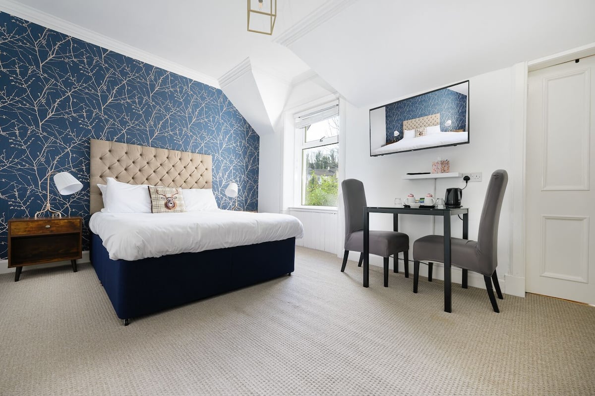 A bedroom with a large bed, a desk with chairs, a window, and a wall with a tree pattern.