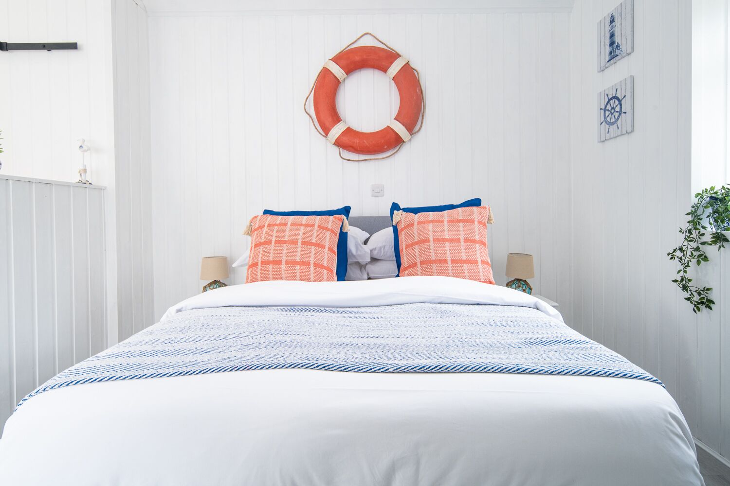 Una cama bien hecha con una colcha blanca y azul estampada, dos almohadas con detalles naranja y azul, un flotador colgado en la pared y un reloj pequeño.