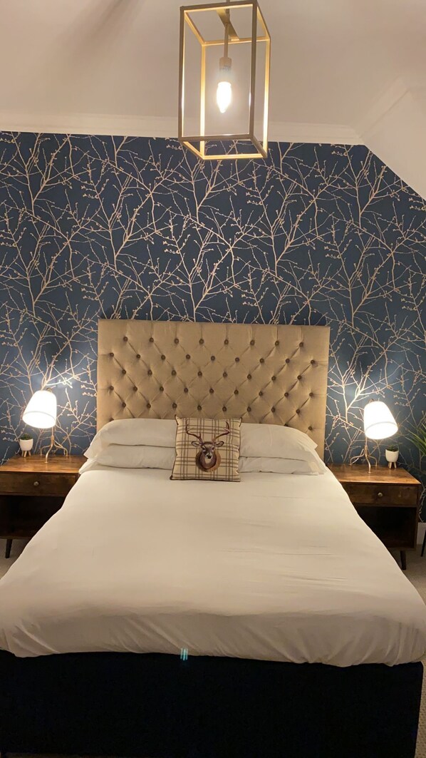 Un dormitorio con cabecera capitonada, una cama grande, dos lámparas de noche y una pared con estampado de ramas de árbol.