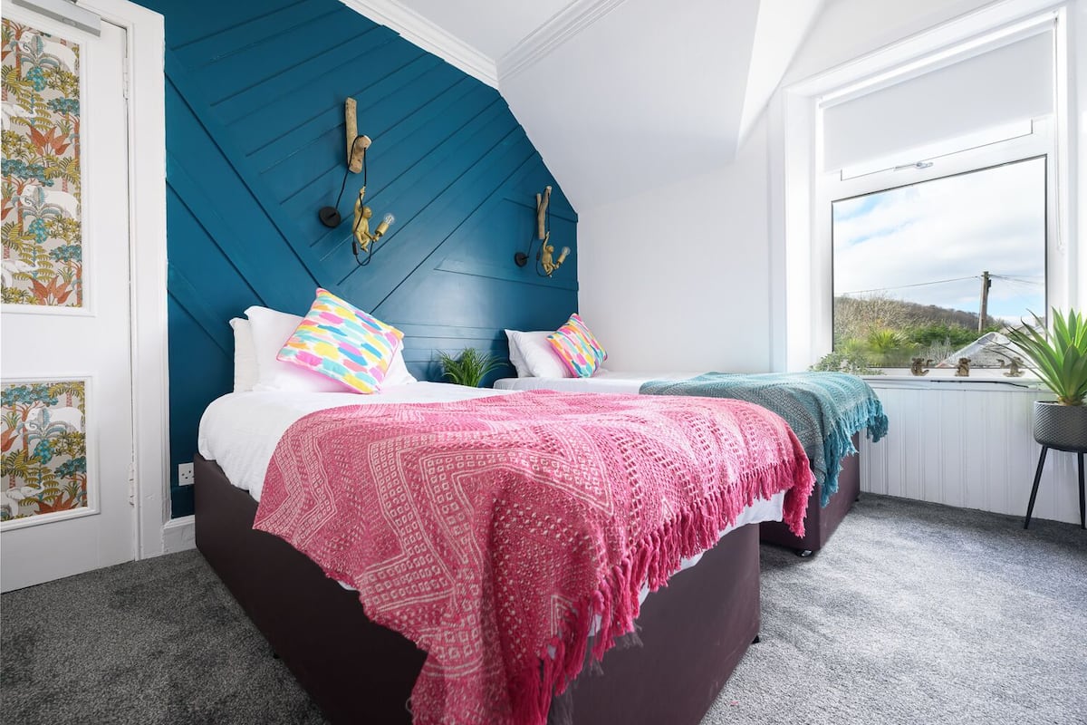 A bedroom with a dark brown bed, two pillows, a pink blanket, and a teal blanket. There is a window with a view of greenery and a plant on a stand.