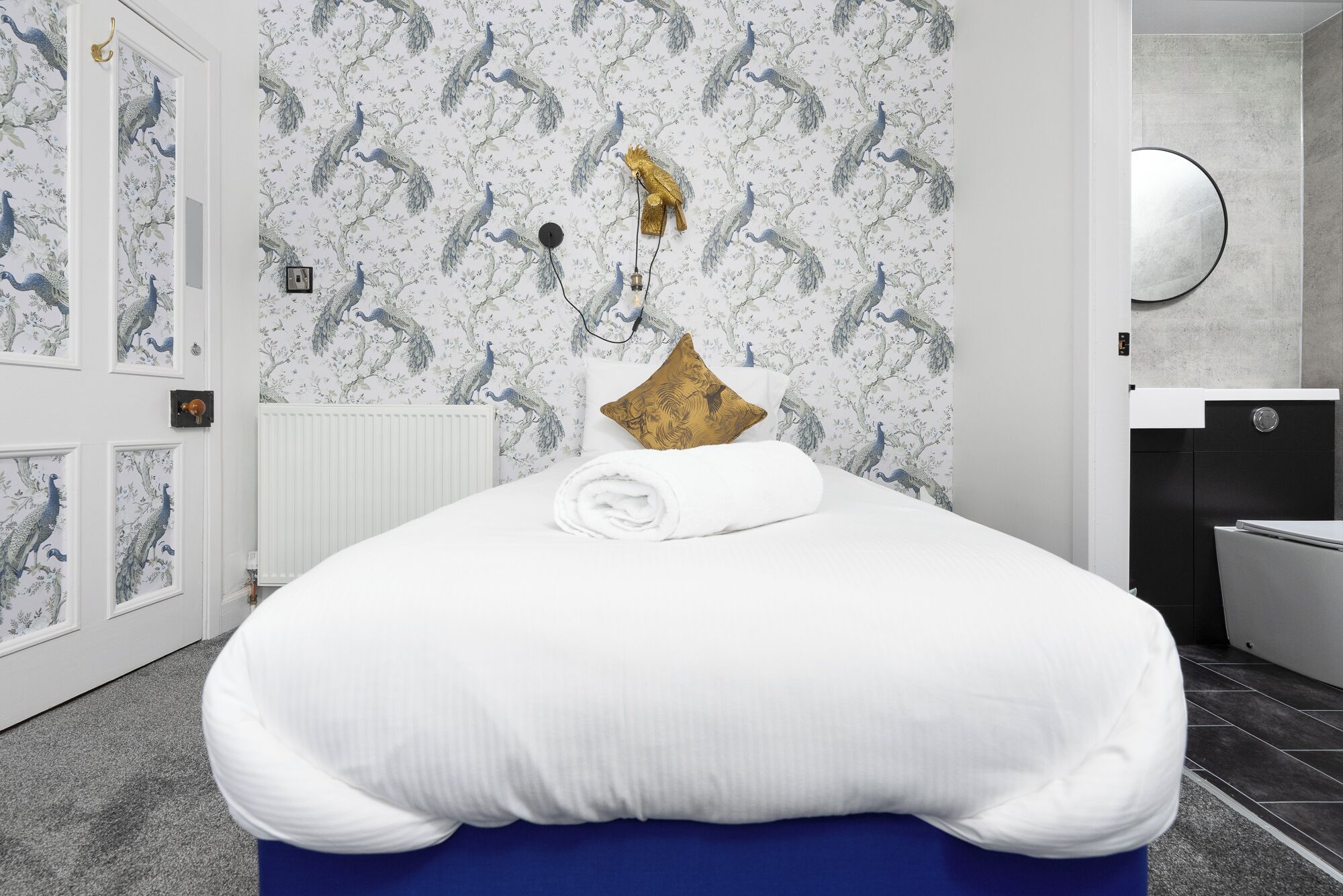 A bedroom with a bed, a radiator, a wall with peacock wallpaper, and a bathroom with a toilet and sink.