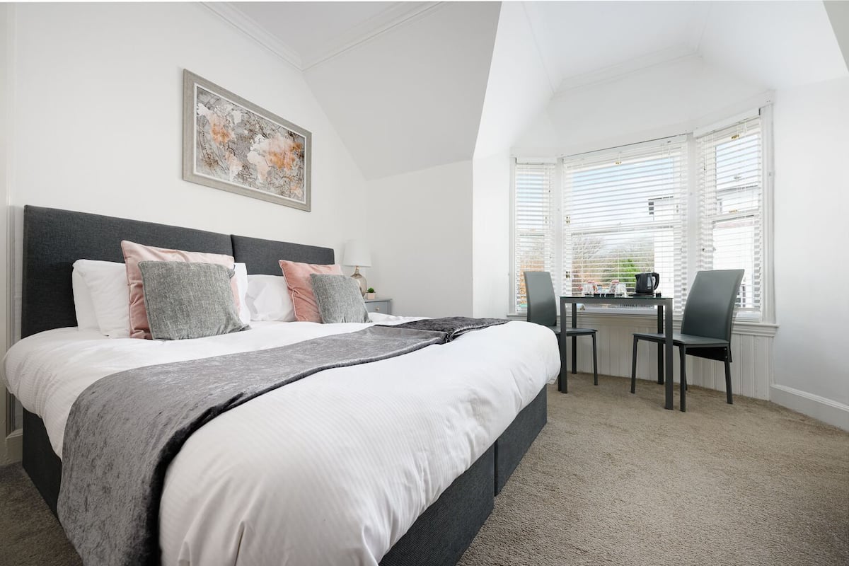 A bedroom with a bed, a desk, and chairs, a window with blinds, and a painting on the wall.