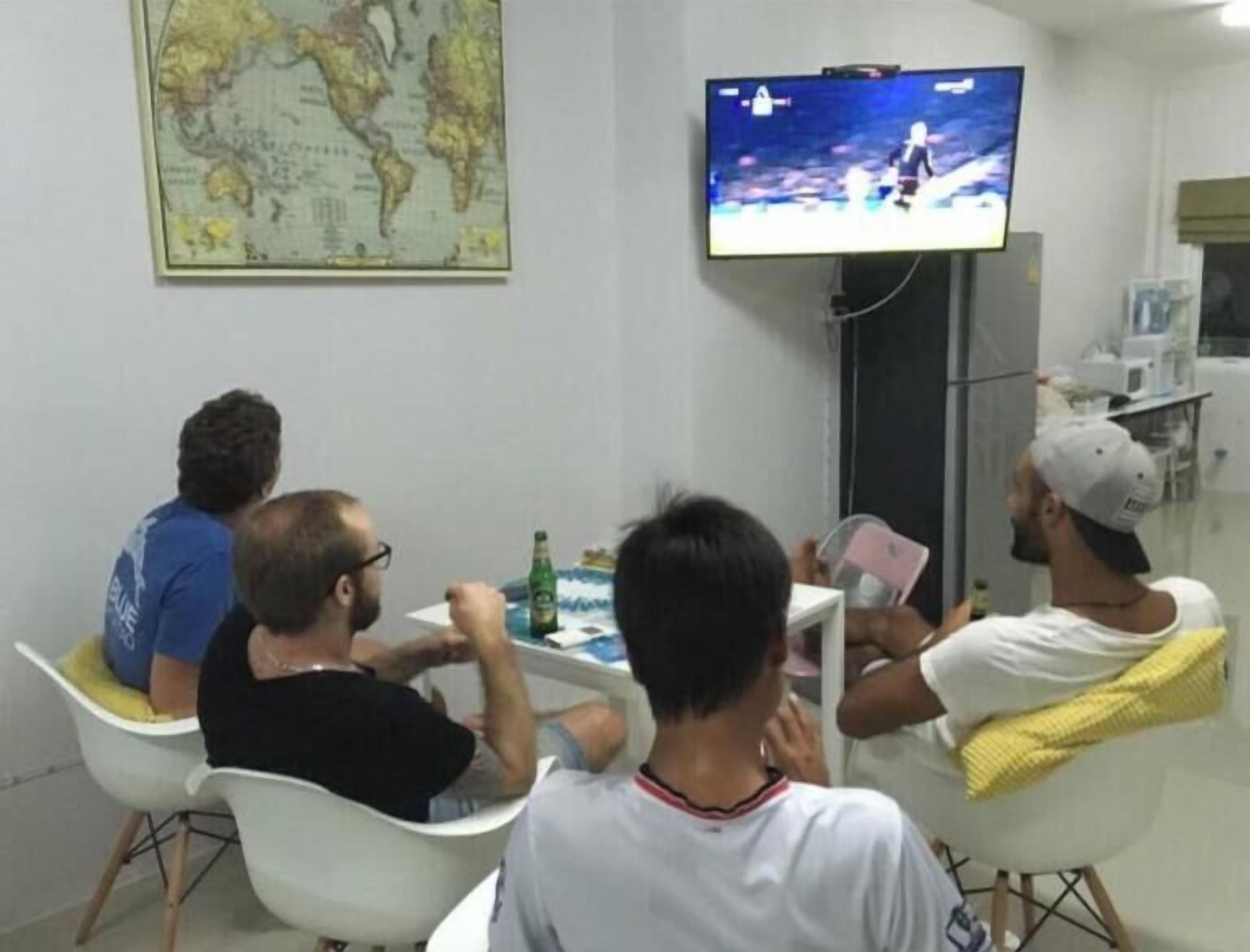 A group of people seated around a table with a television displaying a video game, in a room with a world map on the wall.