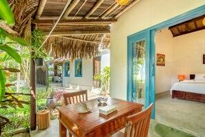 A wooden dining table with chairs, a thatched roof, and a view of a bedroom with a bed and a nightstand. - Ocean House (Da Nang)