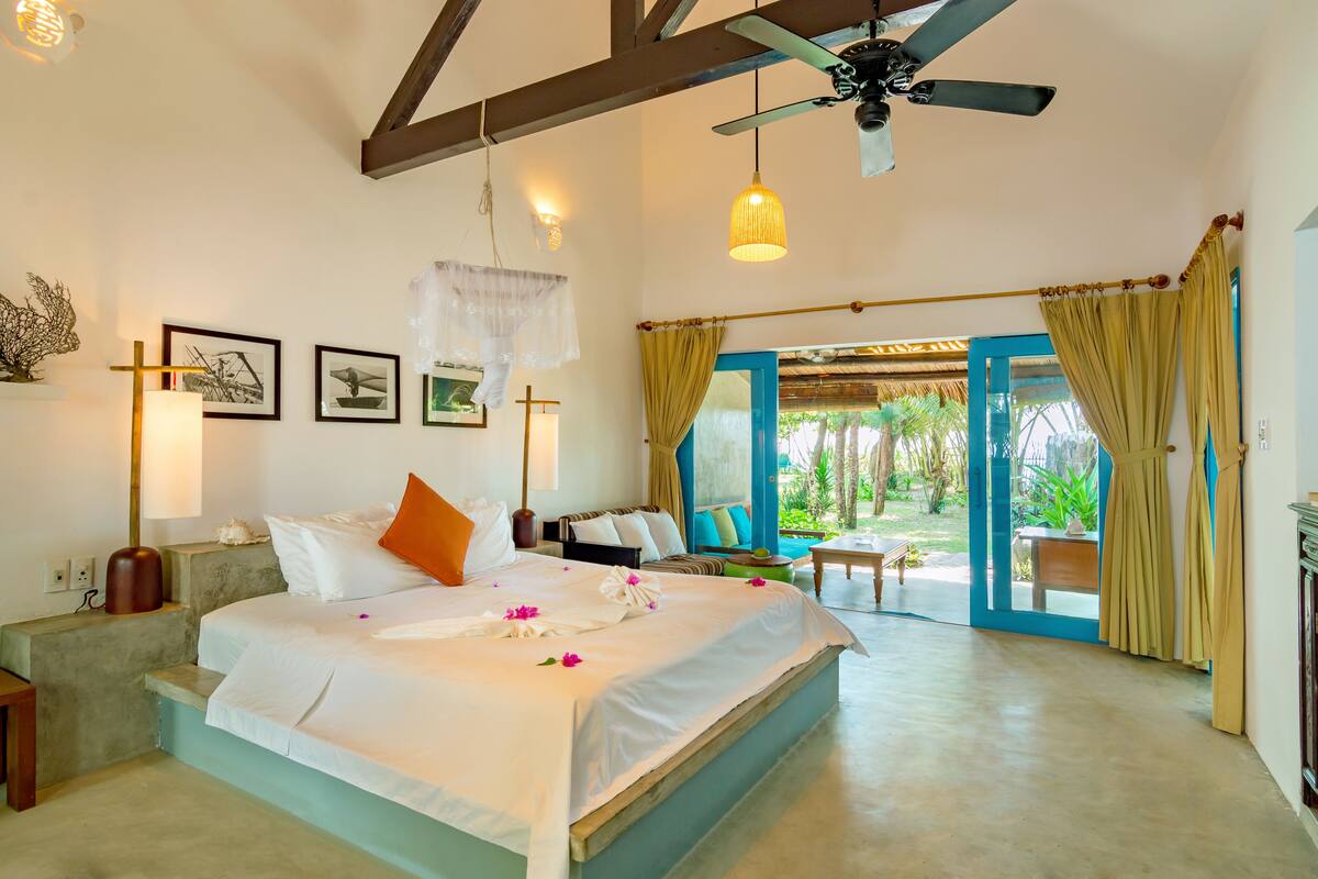A spacious bedroom with a large bed, a ceiling fan, and a view of the garden through an open door.