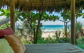 A beach view from a thatched-roof hut with a bed and pillows, overlooking a sandy beach with umbrellas.