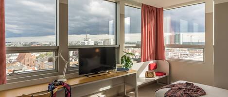 A modern hotel room with a large window, a desk with a computer, a bed, and a chair.